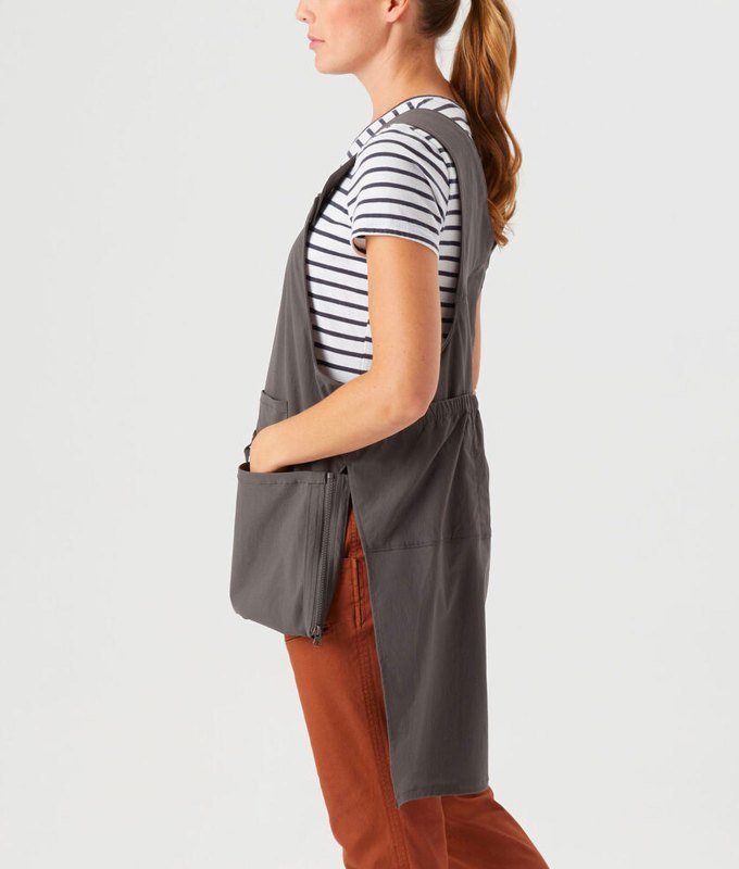 best-gardening-gifts: A woman wears an asymmetrical gardening apron that is longer in the back and shorter in the front with pockets for tools. She also wears a pair of terracotta colored pants and a black and white striped shirt in front of a gray background.