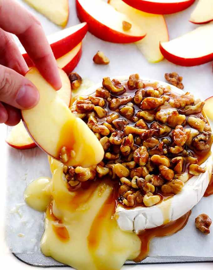 fun things to bake: baked brie topped with caramel and nuts, with a hand dipping an apple into the oozing cheese