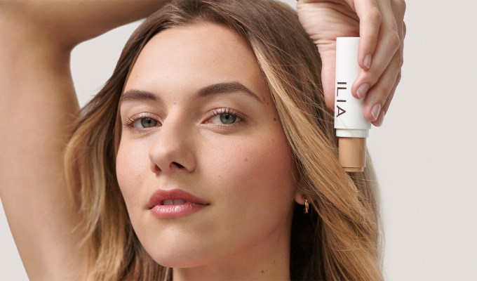 A woman holding up the new ILIA Skin Rewind Complexion Stick next to her face.