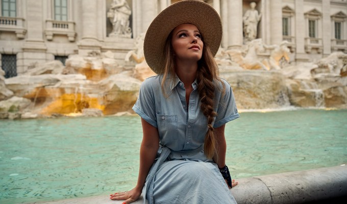 poses for pictures: woman wearing dress and hat sits in front of trevi fountain in rome