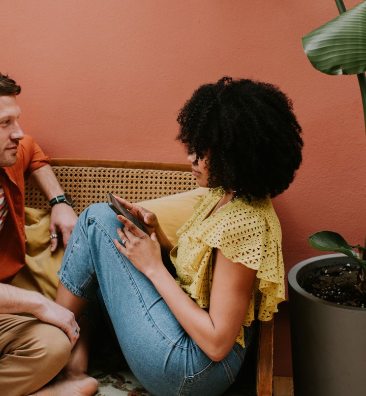 questions-for-couples: A beautiful couple sit on a rattan seat in an outdoor area against a pink wall. The man turns in towards his partner, and looks at her. She sits with her legs up on the seat, against him, and looks back.